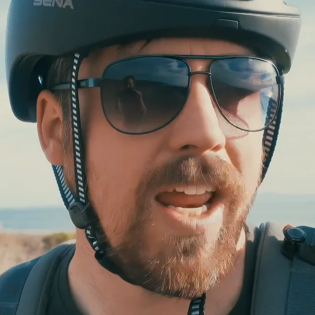 Andy Robinson in a helmet and aviator sunglasses, an action camera on his chest strap, talking to camera by the highway.