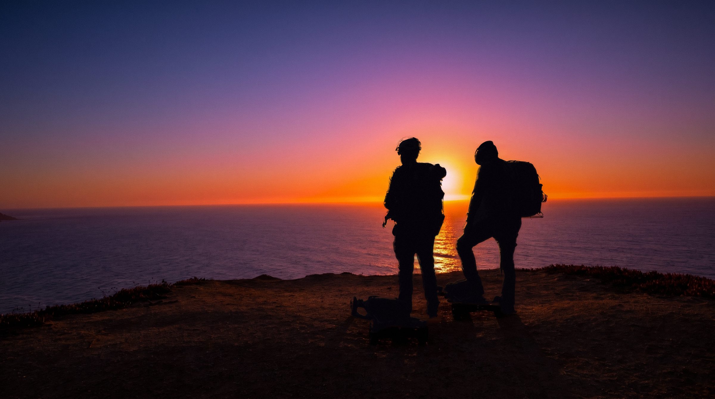 The two riders in silhouette on a clifftop above the Pacific at sunset, their boards at their feet.