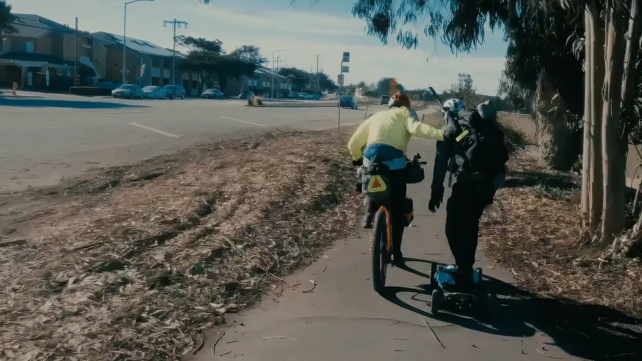 A rider with a dead board being towed along the bike path by a touring cyclist in a yellow jacket.