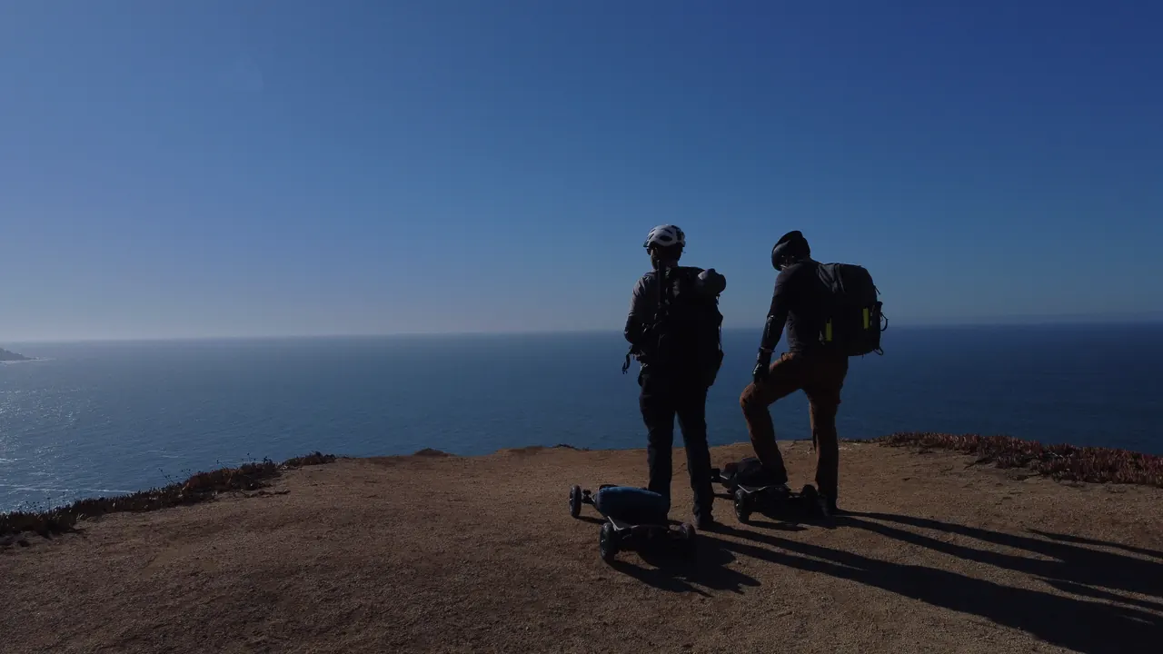From a drone: the two riders in silhouette on a clifftop above the Pacific, boards at their feet.