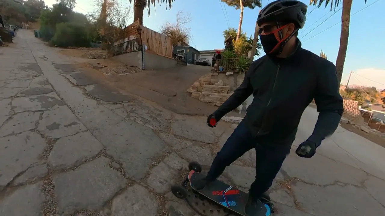 Andy, masked and suited up, riding the board up a steep, cracked residential street.