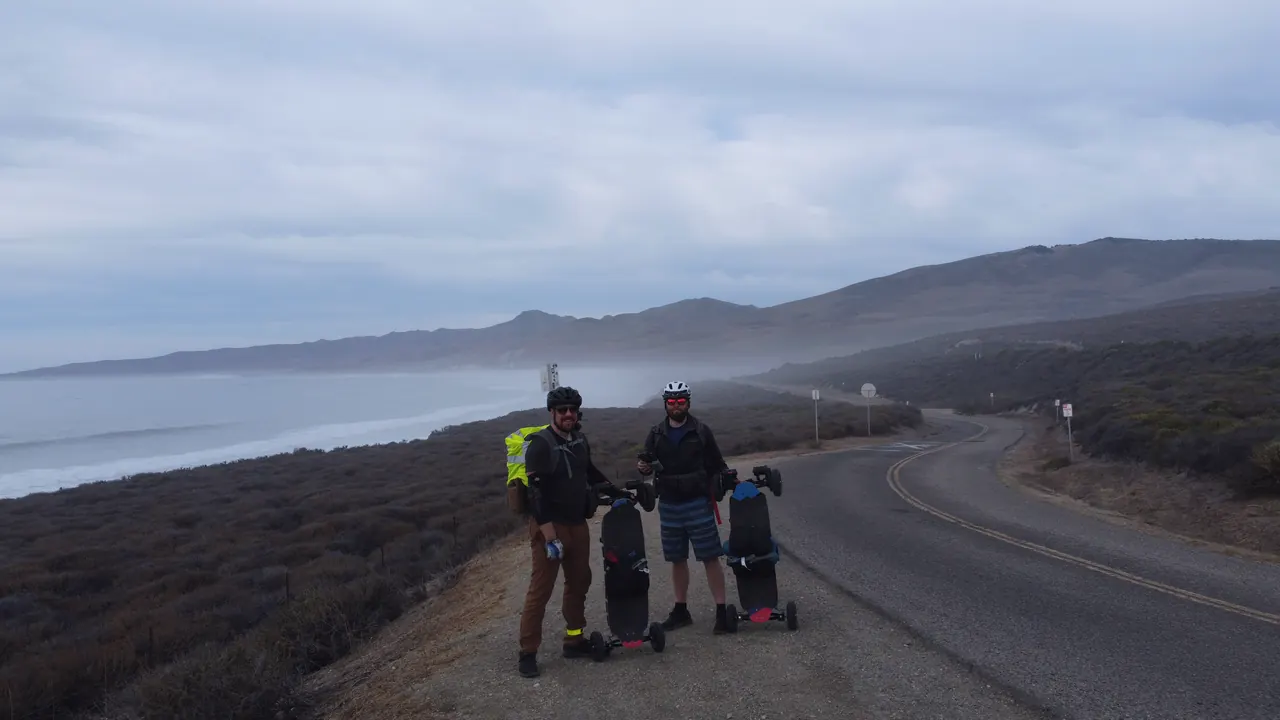 From a drone: the two riders on the coast road to Jalama Beach, holding their boards upright.