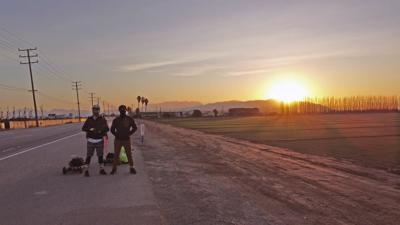 From a drone: the two riders standing on the road at sunrise, the sun coming up behind the mountains.