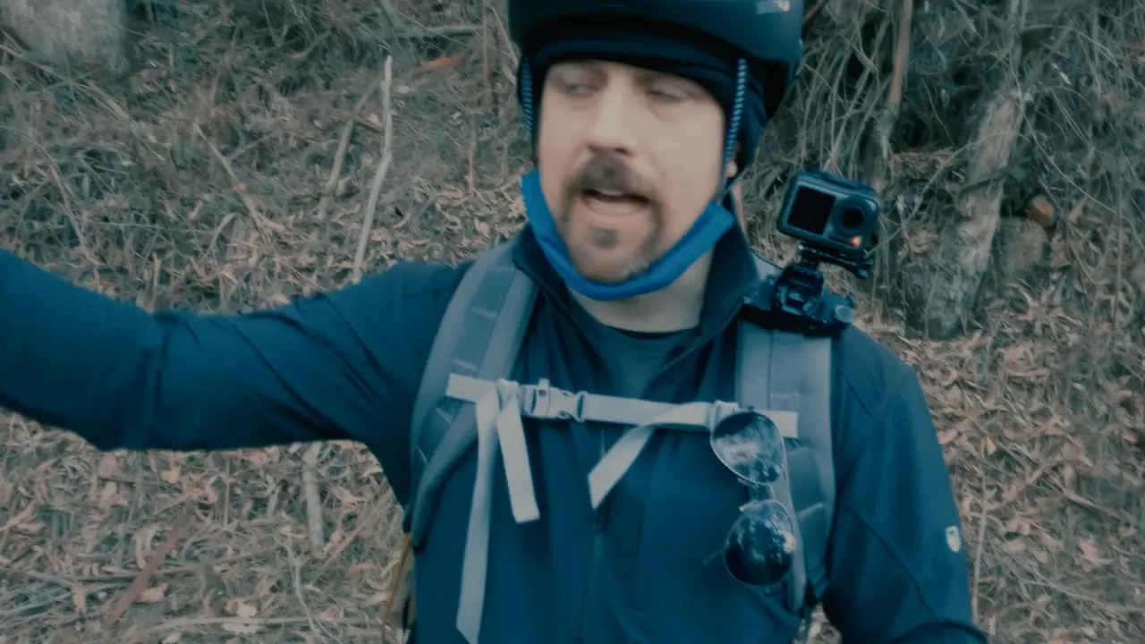 A rider in a helmet and backpack by the roadside, pointing at the traffic.